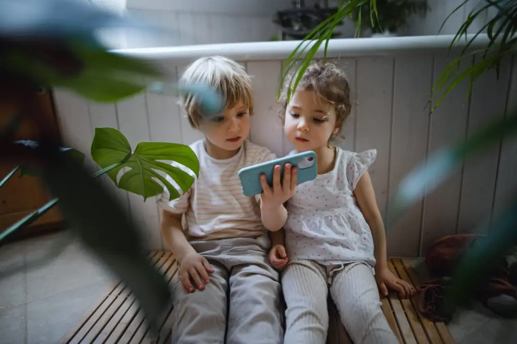 Cute small children sitting on floor indoors at bathroom, using smartphone